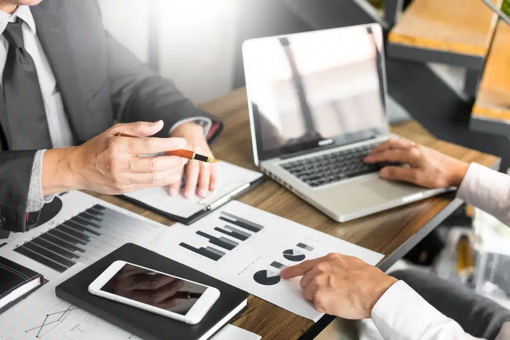 mesa de escritório com um notebook, papeis, um celular, além de duas pessoas prestando serviços contábeis